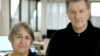 Ein Foto von Anne Lacaton und Jean Philippe Vassal, beide in Schwarz gekleidet mit kurzen grauen Haaren und zurückhaltendem Lächeln. Im Hintergrund sind große helle Fenster und weiße transparente Vorhänge.
