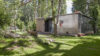 Foto vom Migrantpolitan auf Kampnagel. Ein kleines einstöckiges Haus mit einem grünen Garten, Bänken aus Holzpaletten, umsäumt von vielen Bäumen.