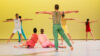 5 Tänzer*innen in bunten Kostümen stehen mit ausgestreckten Armen vor einer gelben Wand. Zwei Tänzer*innen in rosa und roten Kostümen sitzen in der Mitte des Raumes aneinander gelehnt auf dem weißen Boden.