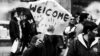 Black and white photo of a protest. One person is carrying a sign with two handprints and the word Welcome painted on it.