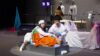 Auf einer Bühne liegt eine Person in orangener Kleidung mit schmerzverzerrtem Gesicht auf einem Podest, zwei Personen in weißer Kleidung bearbeiten sie, halten die Hand und messen Puls. Hinten stehen zwei Männer in weißer Uniform, mit Pappbechern im Mund.