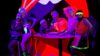 Sechs Personen in Sportkleidung im 80er-Jahre Stil posieren um einen Tresen in lila und rotem Neonlicht herum. Sie tragen fluoreszierendes Make-Up, im Hintergrund ist ein ca. 3 Meter großer knallroter Mund mit offenen Lippen und zwei Schneidezähnen.