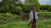 Ron Sexsmith steht in einer grünen Landschaft und hält seine Gitarre in der Hand.