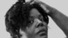 Ein schwarz-weiß Foto von Mable Preach: Eine Schwarze Frau mit kurzen, welligen Haaren. Sie trägt ein schlichtes T-Shirt und silberne Ohrringe. Sie blickt entspannt aus dem Augenwinkel in die Kamera und hält eine Hand an den Kopf.