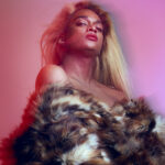 Lotic looks sensually, slightly from above into the camera, wearing red lipstick and an off-the-shoulder fur coat, all against a pink background.