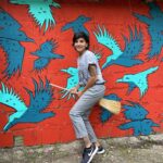 A smiling woman in a grey t-shirt and jeans stands with a broom between her legs in front of a wall on which blue birds are painted. She holds the broom as if she were flying away with it like a witch.