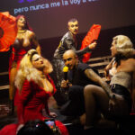 Five performers, some in drag, stand, kneel or sit on a stage with stairs and pose exalted. Two have huge red fans in their hands, one in the middle sings into a yellow microphone.