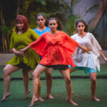 Four women of Colour stand in front of and next to each other with alert gazes. They wear colourful loose dresses made of light fabrics and stand barefoot and wide-legged, each with one leg on tiptoe on artificial grass. The one in front touches the other
