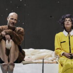 Ein Schauspieler mit Glatze und braunem Wollpulli sitzt mit angewinkelten, nackten Beinen auf einer weißen Plattform. Neben ihm eine Schauspielerin mit dunklen Locken in einem gelben, adretten Kostüm aus Rock und Jackett, die genervt nach oben blickt.
