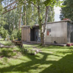 Photo of the Migrantpolitan at Kampnagel. A small one-storey house with a green garden, benches made of wooden pallets, surrounded by many trees.
