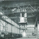 Ein altes schwarz-weiß Foto der ehemaligen Fabrikhallen von Kampnagel mit Gerüsten, einem Kran und aufgereihten Metall-Tonnen.