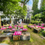 Kampnagel's Waldbühne (forest stage) under large green trees. In front of it are large stones lined up for sitting with neon pink cushions. On the stage, some people are standing and talking.
