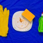 Auf einem knallblauen Tuch liegen ein Paar gelbe Gummihandschuhe, ein schmutziger weißer Porzellanteller mit einem gelben Schwamm und eine grüne Spülmittelflasche.