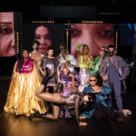 Ten colourfully dressed performers stand and kneel on a stage with serious looks. Behind them hang screens with close-ups of faces and a table with a sound system stands at the right edge.