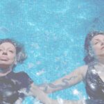 Little Annie and Evilyn Frantic are swimming on their backs in a light blue pool, they are both wearing black shirts, only their faces are above the water surface.