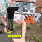 Eine Hand mit einem Smartphone zeigt auf einen QR-Code auf den Boden mit Graffitis an einer Mauerwand im Hintergrund.
