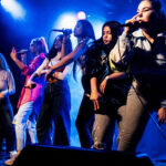 6 young women with long straight hair stand on a stage and perform with microphones in their hands. They stand dynamically in a row and wear colourful clothes. From behind blue lights are shining on them