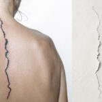 Auf dem Rücken einer weißen Person ist eine schwarze unregelmäßige Linie entlang der Wirbelsäule tattoowiert. Rechts daneben ist die Vorlage des Tattoos: Ein Riss in der Wand.