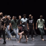 Viele Tänzer*innen stehen in verschiedenen Positionen auf einer Bühne mit schwarzem Hintergrund. Eine Person macht eine Break-Dance Bewegung auf dem Boden. Die Tänzer*innen schauen diese Person an.