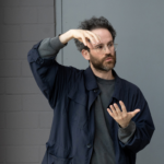 Eine männlich gelesene Person in einem grauen Pullover und schwarzen Hemd hält beide Hände in einer Haltung, als würden sie eine Kugel halten.