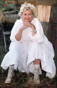 Eine weiße Person mittleren Alters sitzt, mit dem Kinn auf ihre Handrücken gestützt, in einem langen weißen Kleid zwischen Baumstümpfen im Gras und schaut lächelnd in die Kamera.