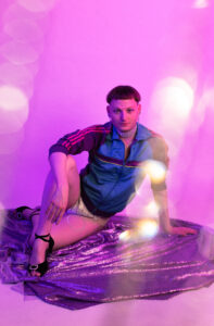 Photo of a white person with a round face and a mushroom haircut, sitting on a cloth on the floor, one arm resting on a bent knee. The person is wearing a blue track jacket and silver glittery shorts and black strappy pumps.