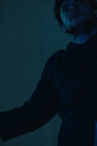 Teile des Gesichts und des Oberkörpers eines Tänzers mit kurzem dunklen Haar und Schnurrbart, in blaues Licht getaucht.