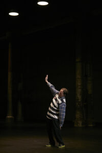 A performer in a plaid shirt and striped sweater stands in a black stage space and stretches a hand up into the air; tree trunks can be glimpsed behind him.