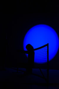 In front of a deep blue light cone, the black silhouettes of a badminton net and a standing performer can be seen in profile.