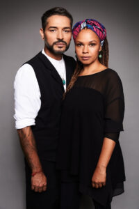 Gianni Jovanovic and Oyindamola Alashe stand close together against a gray background. He in a white shirt and black vest, she in a black dress and colorful hair band, her long, braided hair falling over her shoulder.
