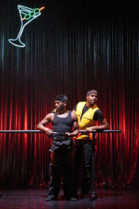 Zwei Schwarze Männer stehen an einer Ballettstange vor einem rot leuchteten Vorhang. Sie tragen schwere Hosen und schwarze, enge Shirts sowie schwarze Durags auf dem Kopf. Einer trägt eine gelbe Warnweste. Im Hintergrund hängt ein Neon-Cocktailglas-Schild