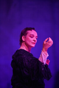 A performer in a black costume stands in front of a purple wall and is lit in pink. She has very short hair and is looking at her right wrist, which she holds in front of her face.