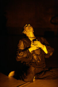 A performer in a black costume made of transparent mesh fabric sits on the floor with his knees bent. He tilts his head back and holds his hands in front of his heart. He is lit in yellow, the background is very dark.