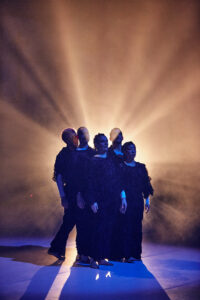 Five performers stand close together in a bluish stage space and are illuminated from behind with warm light. The light appears like rays of sunlight behind them, the stage fog contributes to a sublime atmosphere.
