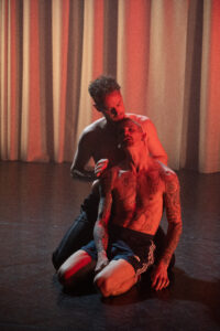 A thin, white, heavily tattooed man, topless and in shorts, kneels on the floor and leans backwards against another white topless man. The man behind touches the man in front tenderly on the head. The light is soft red.