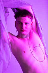 A white topless person with a mushroom haircut and a glittering silver collar leans to the side and stretches their arms above their head. A transparent cloth is wrapped around the arms, a silver squiggly decoration is stuck on the chest.