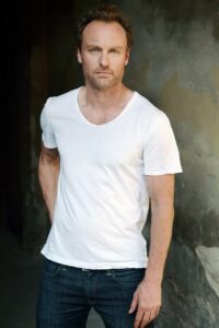 Porträtfoto eines weißen Mannes mit dunklen kurzen Haaren und leichten Geheimratsecken. Er trägt ein weißes T-Shirt und steht mit ernstem Blick und leicht nach vorne gelehnt vor einer Betonwand.