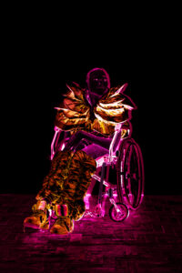 Vor einem tiefschwarzen Hintergrund sitzt eine Person, deren Umrisse und Kleidung in orange und pink leuchten, in einem Rollstuhl. Die Hände sind im Schoß gefaltet und die Person scheint wehmütig in die Ferne zu gucken. Ihre Haut sieht dunkel aus.