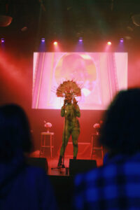A person in a tight full-body suit in high shoes and a fan-like headdress stands on a stage bathed in reddish light and sings passionately. In the foreground there is a blur of spectators.