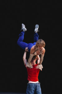 Two well-trained people in front of a deep black background in an acrobatic pose. One person holds the other up with his hands on his hips. The upper person has his legs bent and is leaning on the shoulders of the lower person.