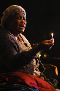 In schummriger Atmosphäre sitzt eine ältere Schwarze Frau in Strickjacke und weicher Mütze. In ihrer rechten Hand ein flammendes Streichholz, das sie klagend anspricht.