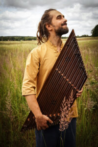 Hakan Tuğrul is standing with his instrument, the Satur, in a meadow with tall grass. He wears a yellowish shirt, a full beard and long hair tied at the nape of his neck and smiles cheerfully to the side.