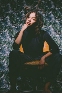 Moyo Ray wears a black tight-fitting turtleneck sweater, short dreads and rests her chin on her hand in a relaxed manner, in the background you can see a floral wallpaper in shades of green and blue.