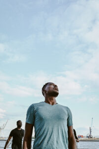A young black man stands in front of a light blue, slightly cloudy sky and looks up. He is wearing a grey-blue T-shirt. Someone else is standing behind him. In the far background, the port of Hamburg and its cranes can be seen.