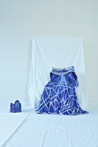 Ein strahlend Blaues Kleid mit weißem Muster liegt auf einem Stuhl drapiert in einem Raum, der mit weißen Stoffen ausgestattet ist. Neben dem Kleid liegt ein Turban aus dem gleichen Stoff auf dem Boden.