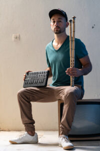 Tayfun Guttstadt is sitting on an old television set with a small mixer and two elongated wind instruments, looking casually into the camera. He wears a dark cap, a green T-shirt and beige trousers.
