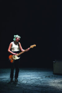Eine junge Person mit grünen Haaren steht singend an einer E-Gitarre auf einer leeren Bühne in dunkelblauem Licht.