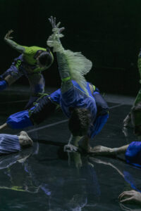Mehrere Personen in blau-grüner langer Kleidung tanzen auf einer dunklen Bühne. Die Person im Zentrum des Bildes steht im angewinckelten Spagat, hat eine Hand auf dem Boden und streckt die andere hoch in die Luft. Sie hat den Kopf zum Boden geneigt.
