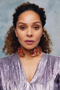 Portrait-Aufnahme von Joy Denalane: Sie trägt ein schimmerndes, lavendelfarbenes Oberteil, ein auffälliges Bronzecollier und ihr Haar im Nacken zusammengebunden. Sie schaut ernst direkt in die Kamera.