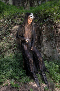 Eine Person in einem Wald trägt ein abstraktes, schwarzes Kostüm mit vielen Falten, das um den Körper zu fließen scheint. Die Person lehnt an einem kleinen matschigen Vorsprung, der von Moss und Geäst bewachsen ist und schaut melancholisch auf den Boden.